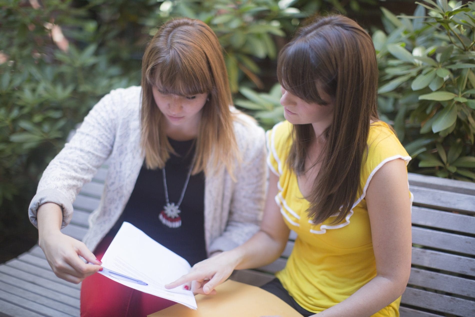 Two women examine a document on a park bench with green bushes behind them.