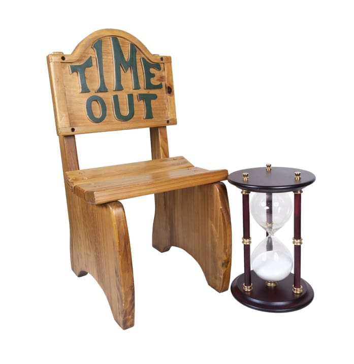 An old-fashioned wooden chair that has the words "Time Out" on it, sitting beside a wood and glass hour glass with the sand in the bottom bulb.